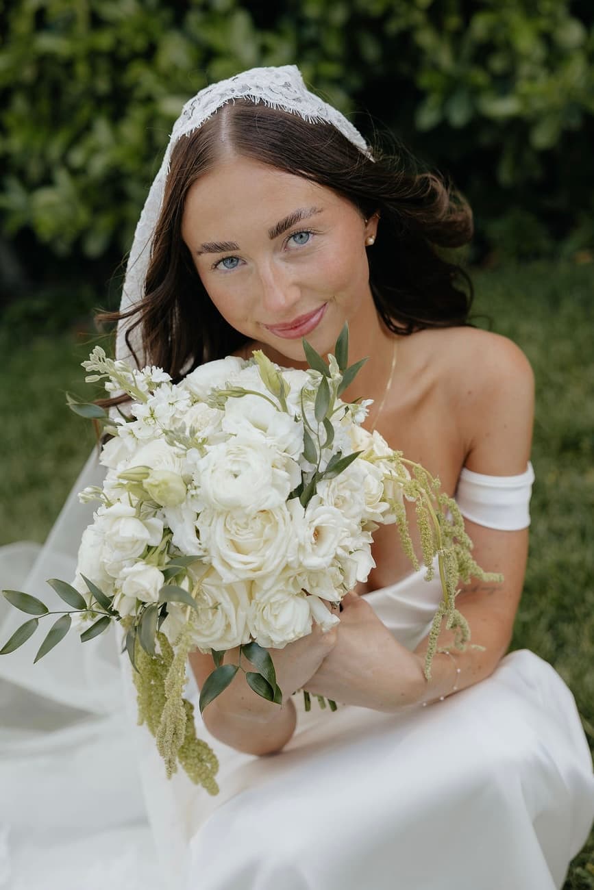 Bride seated on the lawn in a lace veil holding a white rose and ranunculus bouquet with trailing amaranthus