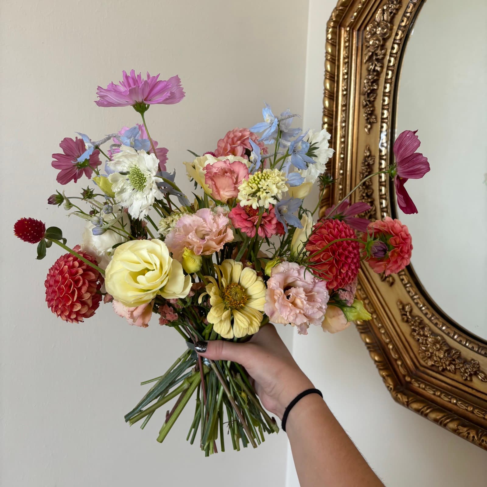 Hand-tied bouquet of dahlias, zinnias, cosmos, and delphinium held up beside a gold gilt mirror