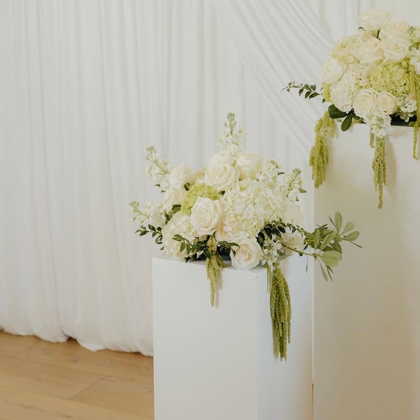 White rose and hydrangea arrangements with trailing amaranthus on white pedestals against a draped backdrop