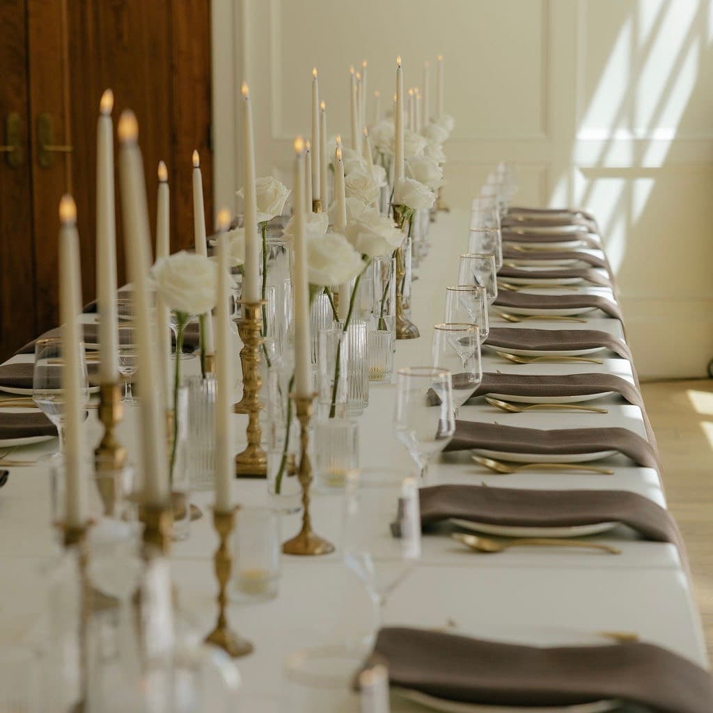 Long reception table set with taper candles, bud vases of white roses, and taupe napkins