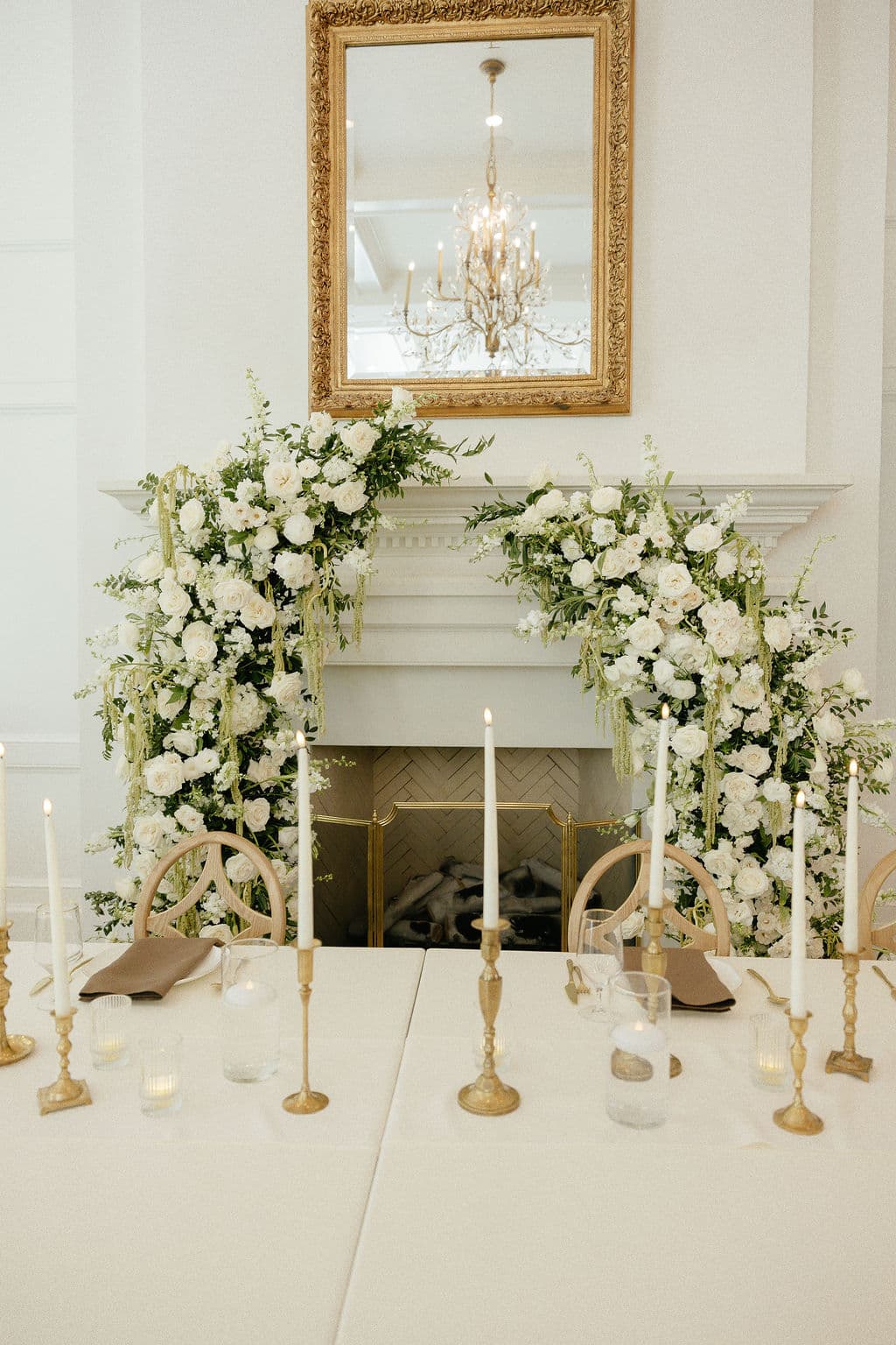 White rose, ranunculus, and stock installations framing a fireplace mantel and gold gilt mirror above a candlelit head table